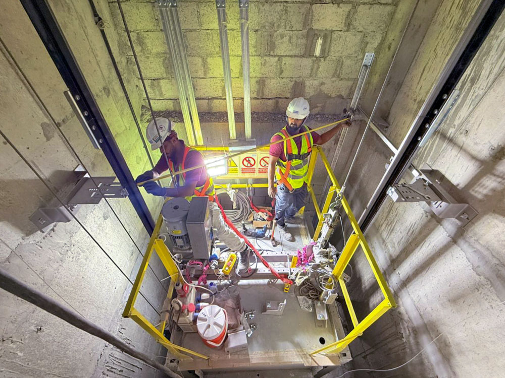 Beach Walk Iii By Imtiaz Two workers in helmets and safety vests are performing maintenance inside a lift shaft with tools and equipment.